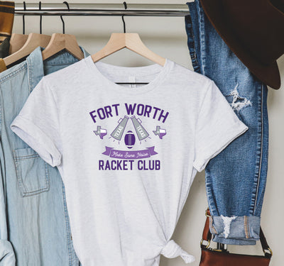 White tee has a graphic that says ""Fort Worth Racket Club" the art also has a banner that says make some noise with a football, two megaphones that say Texas and two Texas Graphics. Colors are purples and grey.